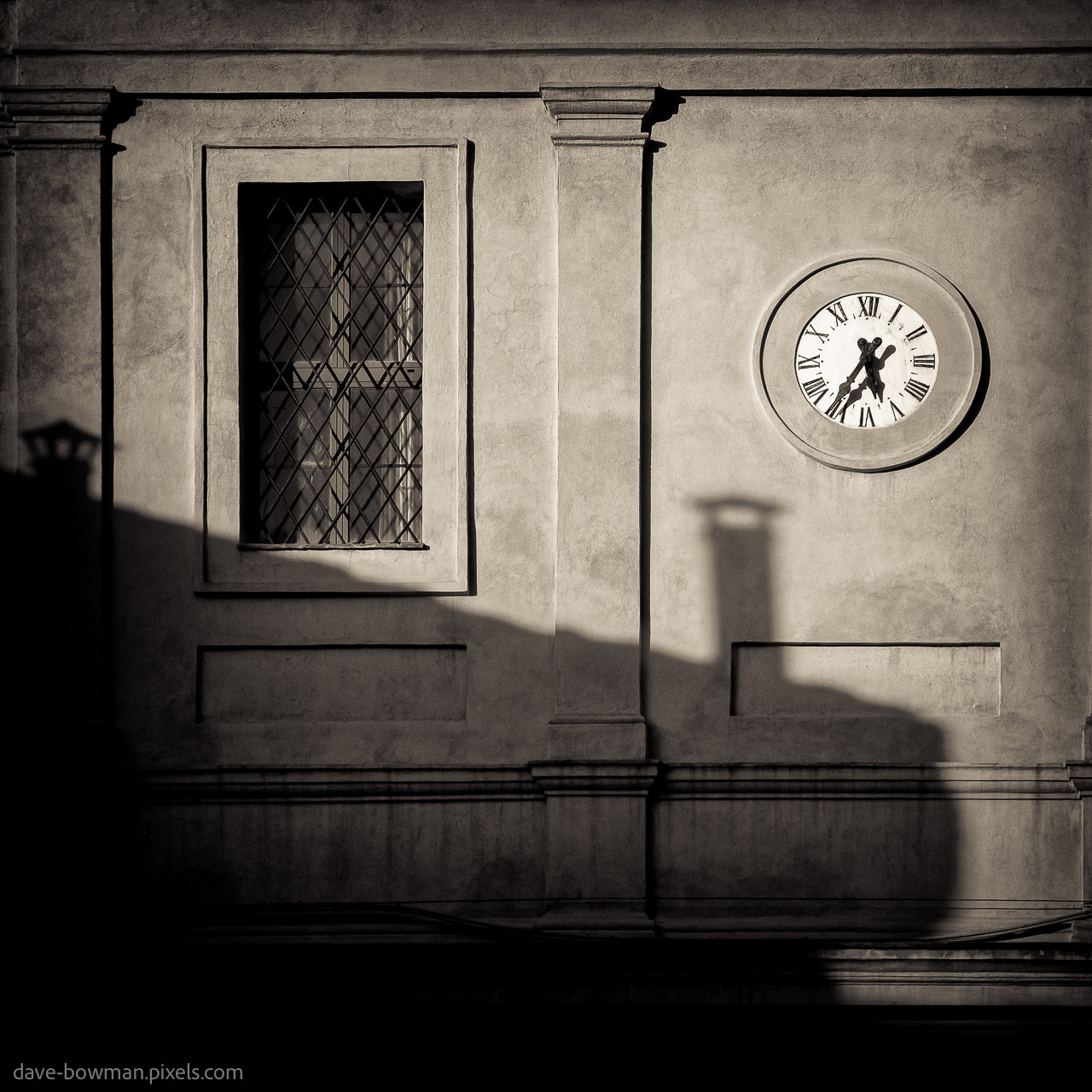 A monochrome, warm-toned photograph of light and shadow on the wall of a building in Siena, Italy. The late afternoon sunlight casts a shadow from an adjacent roof, and there is a white-faced clock embedded in the wall with Roman numerals on it. The clock hands are at 5:35pm. To the left of the clock there is a window.