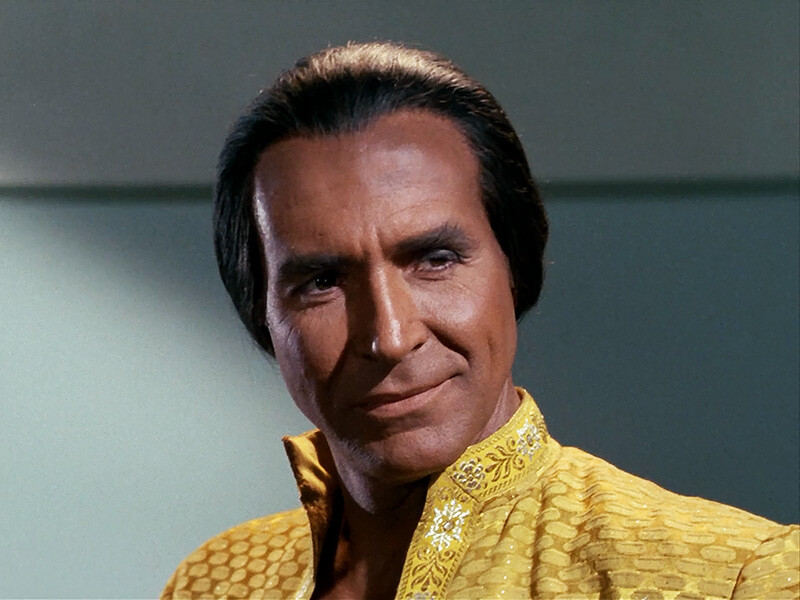 A medium brown skinned man with slicked back brown hair wearing a yellow brocade jacket with a Nehru collar.