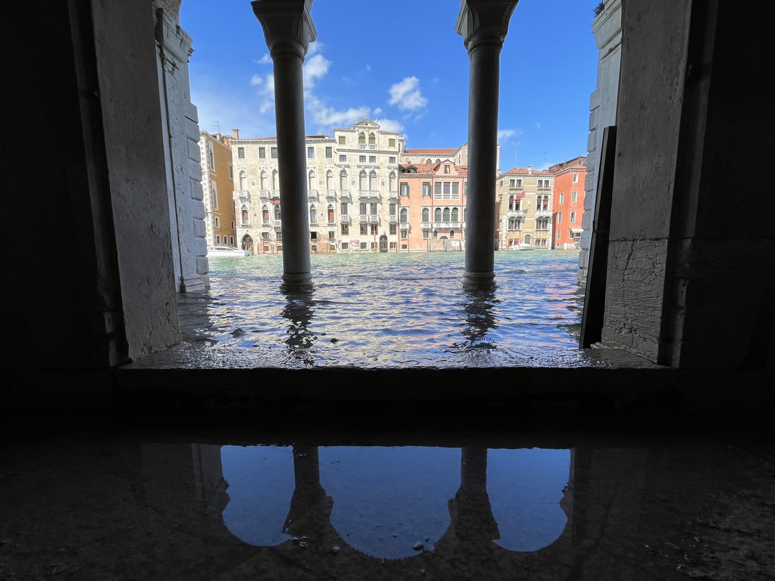 Looking out through a pillared archway onto the Canal Grande. The water has flooded over the edge of the canal and is filling the courtyard in which the photographer is standing, wishing she had put her wellington boots on this morning. The buildings on the other side of the canal and the blue sky above are bright and sunny, in contrast to the dark shadowy pillars. The sky and the top of the archway is reflected in the water on the floor