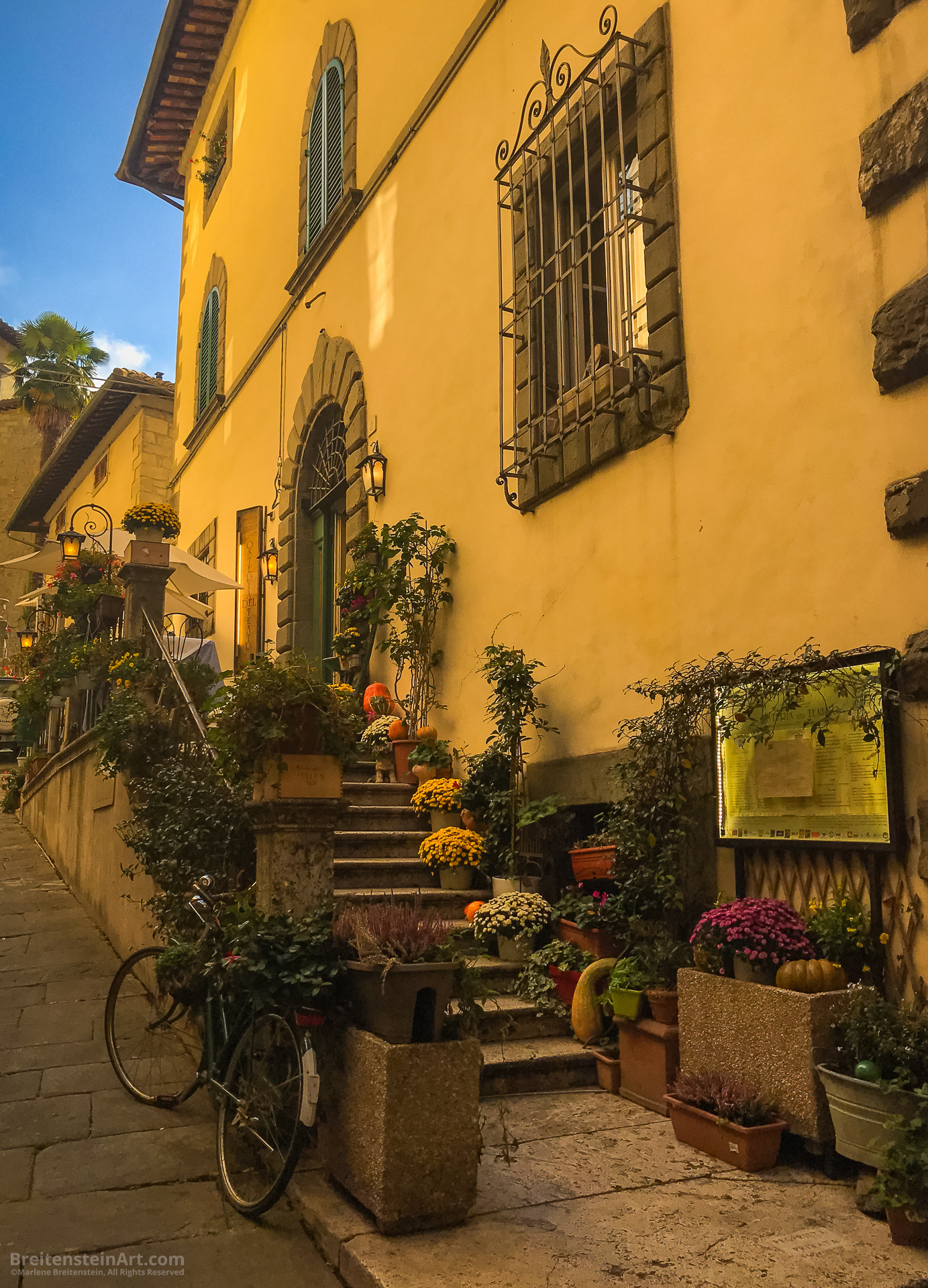 Photograph of a quintessential historic Tuscan street scene, under a sliver of a late-afternoon blue sky. The scene featuring a renaissance-era building with yellow walls—large hewn stones edging the doors and windows—now used as an indoor-outdoor restaurant. Ten or eleven stone steps lead past an illuminated menu, up to a narrow outdoor terrace, with elegant white-clothed tables shaded by wide cream-colored umbrellas. There are many flowering potted plants (in whites, yellows, reds and magentas), creeping vines, and squash, pumpkins and gourds picturesquely lining both sides of the stairs. A leaning bike at the bottom—edging the street’s large cobblestones—is also filled with green plants. Decorative wrought iron grills, with scroll-work designs, adorn a window and the top of the door frame, and warmly lit hanging lanterns are suspended from more curved iron, at the edge of the terrace.