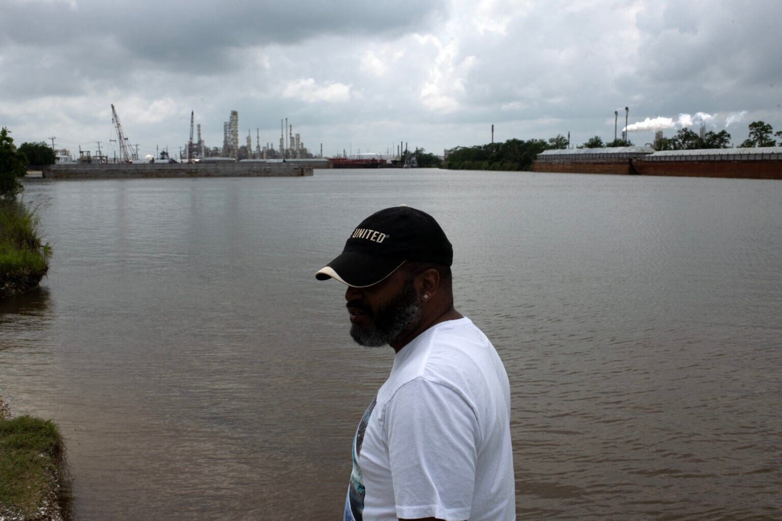 Christopher Jones, a Black man in a baseball cap and white tee, president of the South End Charlton-Pollard Greater Historic Community Association, outside of the ExxonMobil refinery in Beaumont, Texas. The refinery can be seen steaming across part of the Gulf under a cloudy sky. 