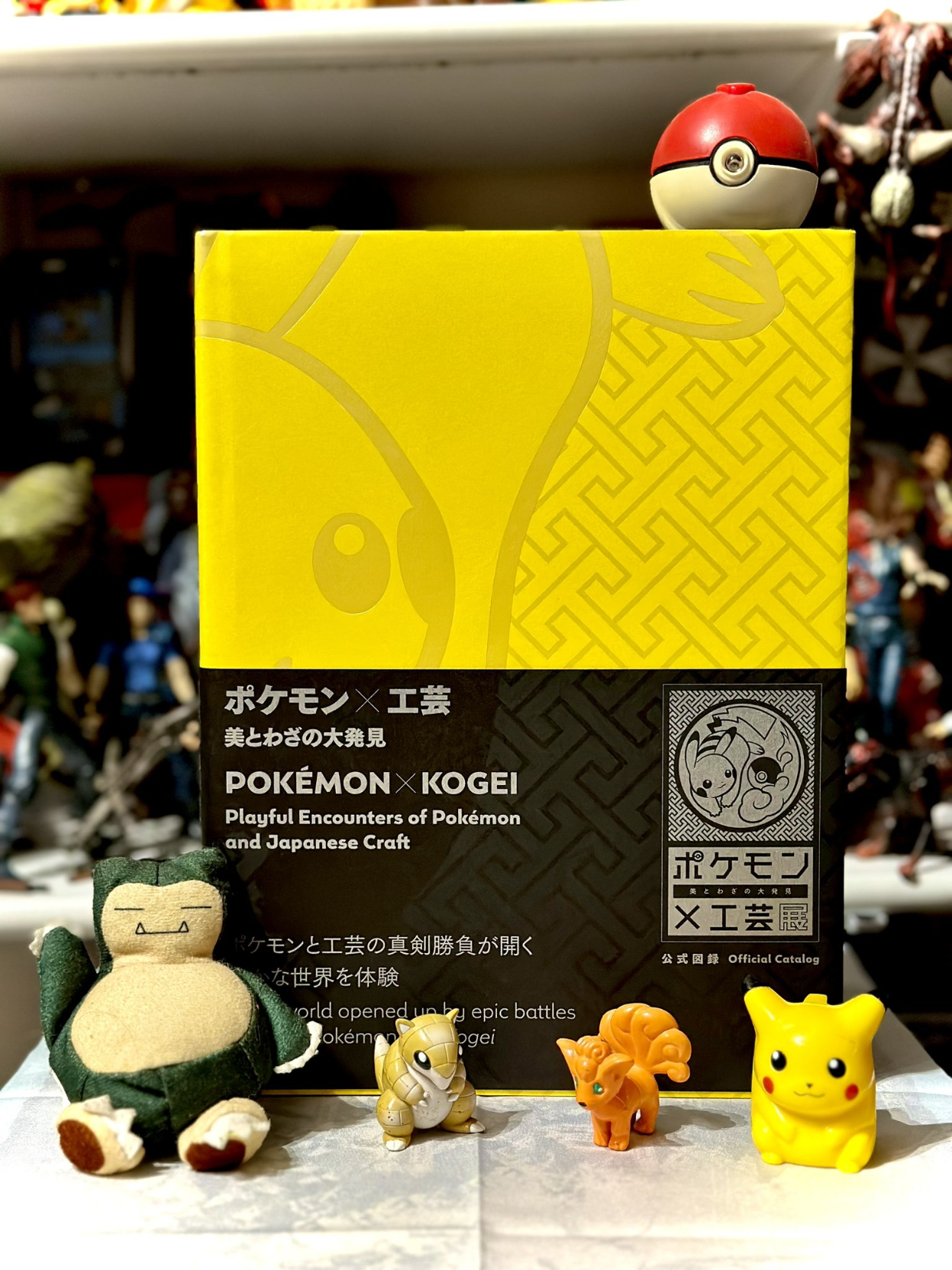 The yellow Pokémon x Kogei book surrounded by Pokémon figurines sitting on a shelf.