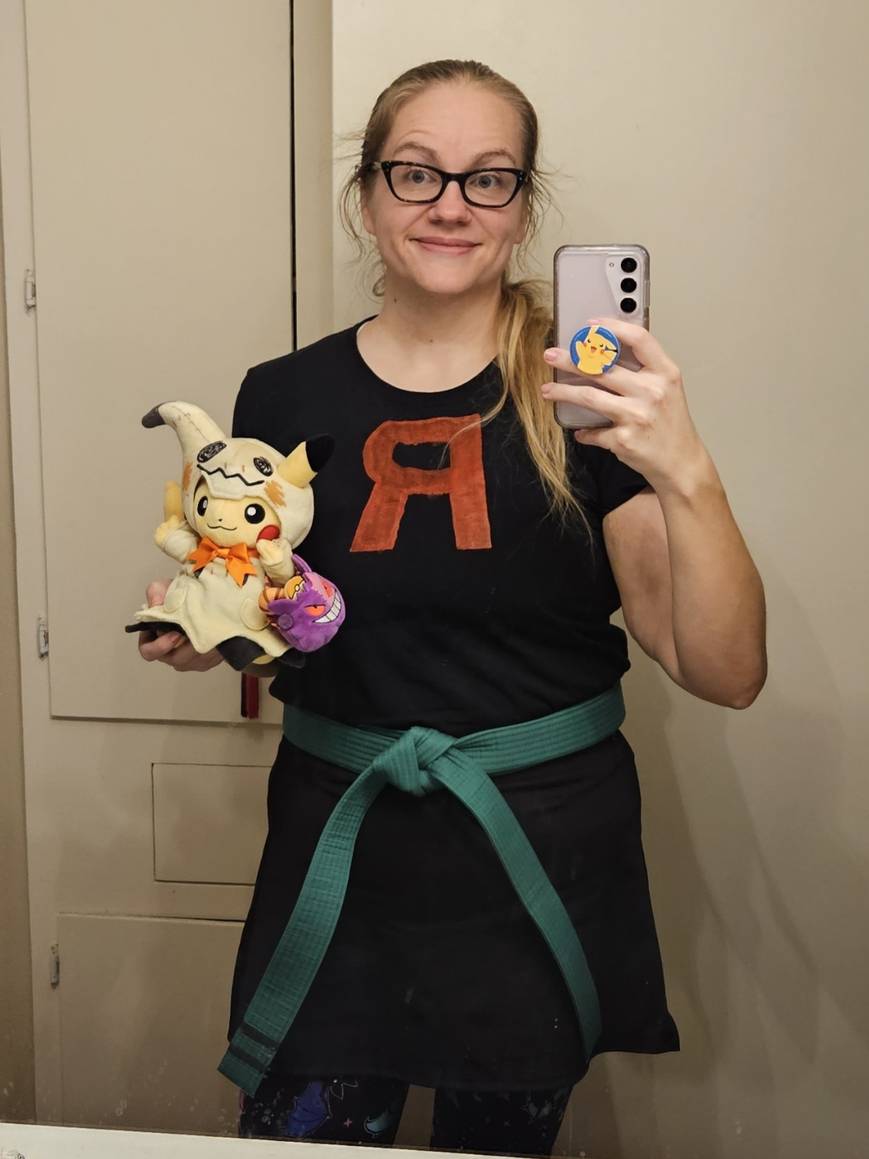 Mirror selfie. I'm wearing a self painted black t-shirt with a large red "R' on it, a black skort, and black leggings with multicolor fluorescent bats on them. I'm holding a Pikachu plush wearing a Mimikyu costume and holding a Gangar shaped Halloween bucket.