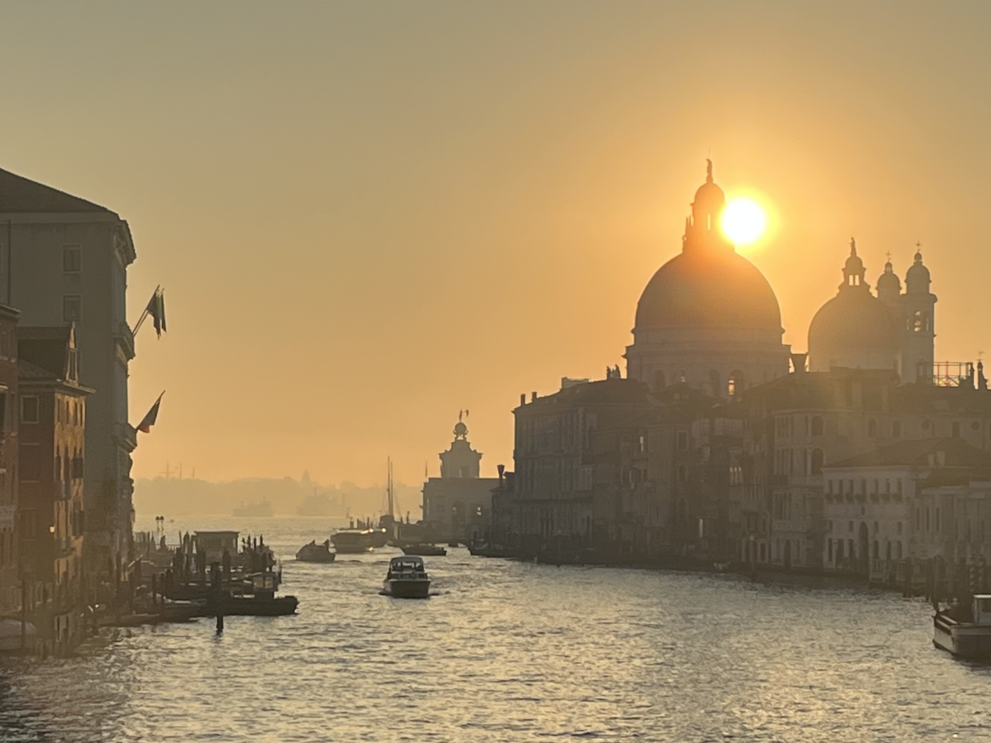 The early morning sun rising through the mist above the dome of Santa Maria della Salute at the mouth of the Canal Grande. The horizon is shrouded in fog, coloured orange by the sun, and the water looks like quicksilver. 