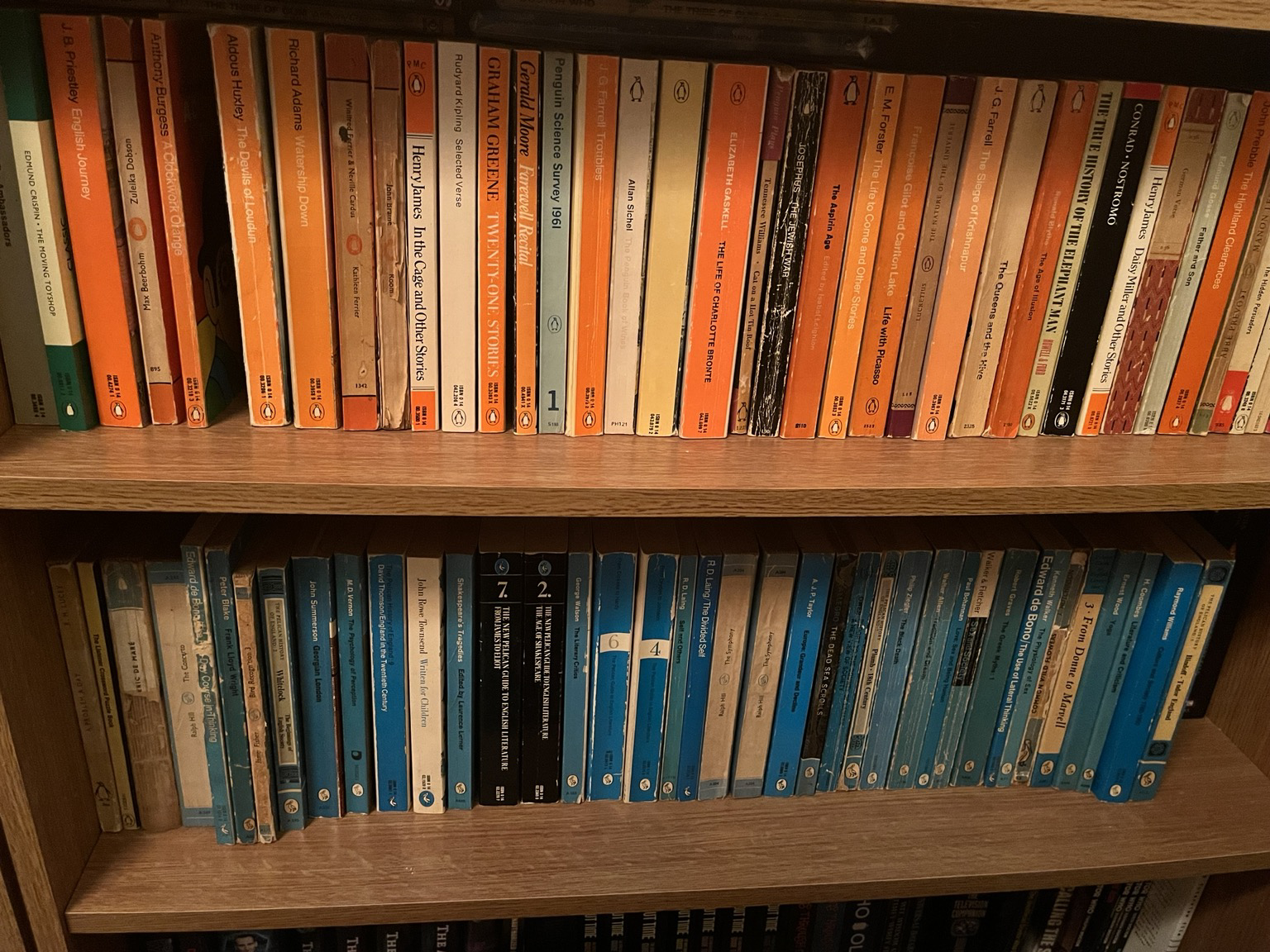 Two shelves of books with the top shelf mostly orange spined Penguin books and the second blue spined Pelican books. 