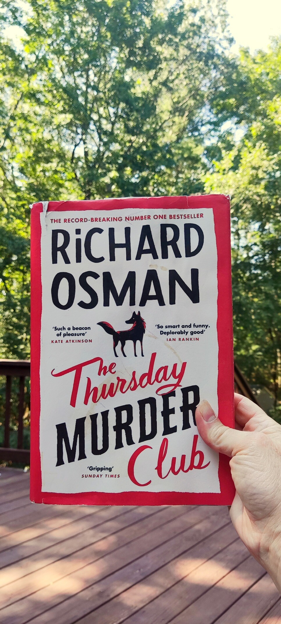 Hand holding THE THURSDAY MURDER CLUB novel against background of trees