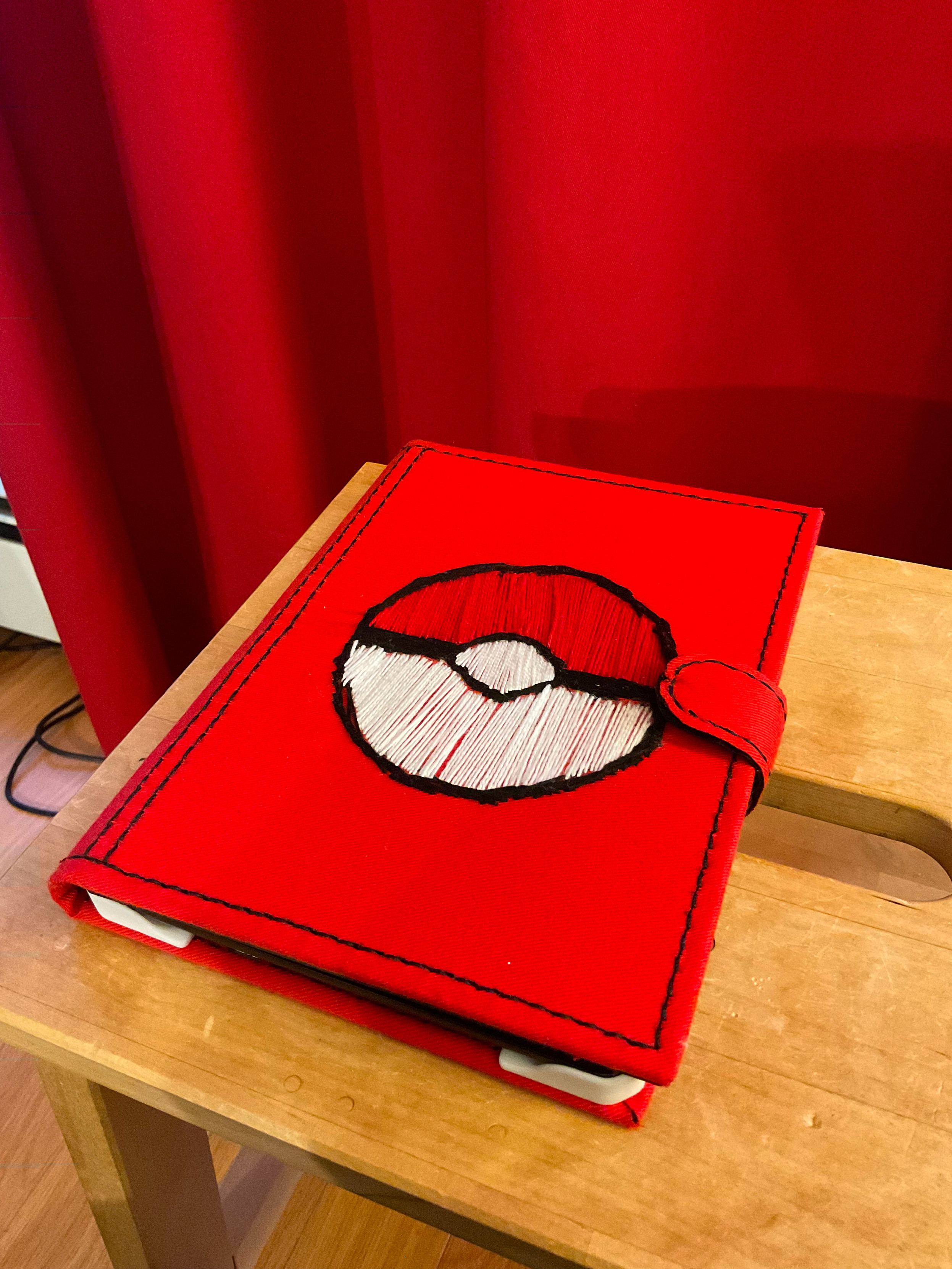 A red e-reader case in cloth with an embroidered Pokeball on the cover.