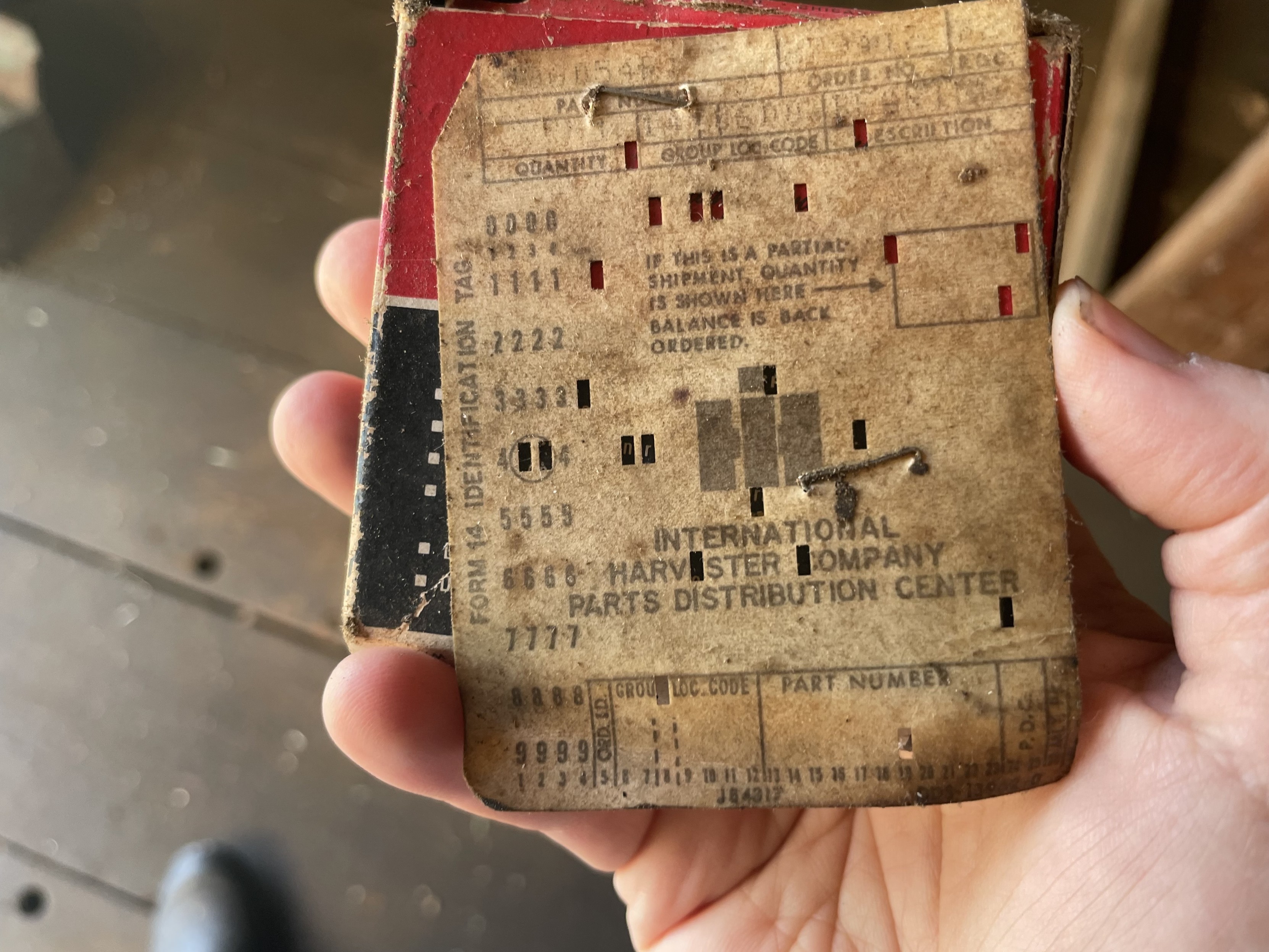 A hand holding a very old red and black box (which contains a larger roller bearing).  Stapled to the box is a half of a very dirty computer punch card that reads International Harvester Parts Distribution Company, and include faded data written on the card about the part.  The punch card has been punched several times.  